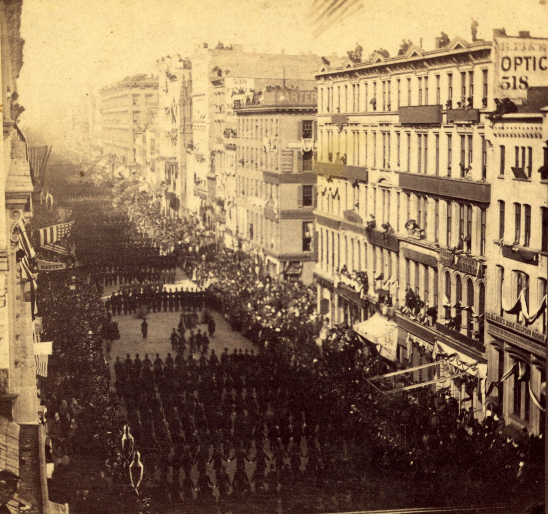 The photograph captures a moment from the funeral procession of President Abraham Lincoln in New York on April 25th, 1865. Crowds of onlookers fill the balconies and windows of the buildings lining the street, while masses of people gather on the sidewalks to witness the event. A large military escort marches in formation ahead of the hearse, which is not visible in the frame, illustrating the grand scale of the public mourning for the assassinated president. The buildings are draped with mourning bunting and American flags, and the solemn mood is palpable even in the still image.