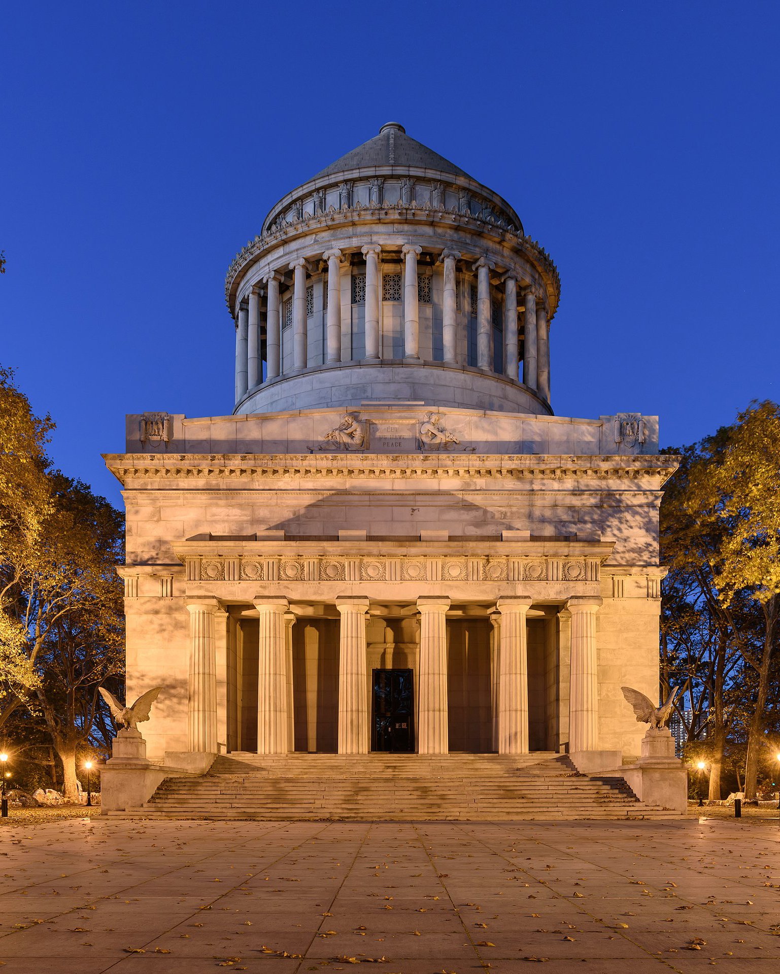 The General Grant National Memorial in Manhattan, New York City, photographed at twilight. The Neoclassical domed mausoleum, also known as Grant's Tomb, is illuminated against the evening sky, with its large granite steps leading up to a portico supported by Ionic columns. Sculptural details and trees in autumn foliage frame the tranquil scene.