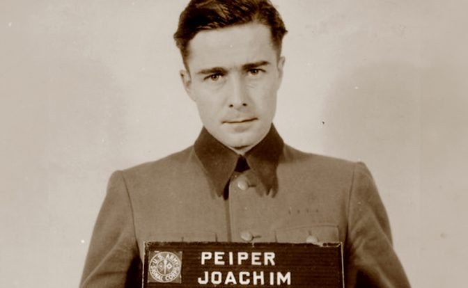 A historical mugshot of Joachim Peiper, taken after his arrest in 1944. He is seen in uniform, holding a nameplate with his name on it, displaying a somber expression.