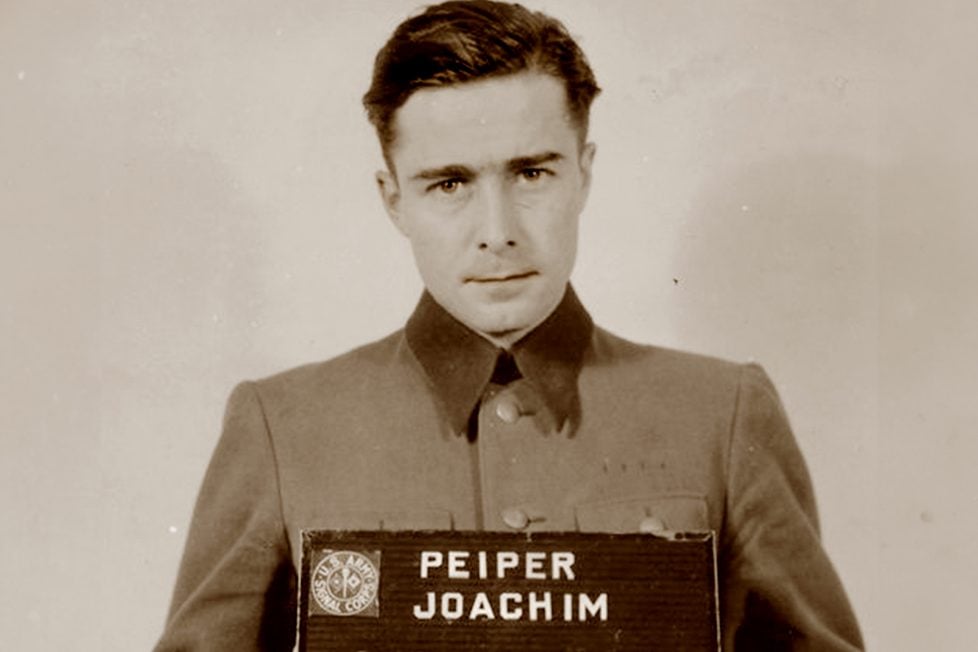 A historical mugshot of Joachim Peiper, taken after his arrest in 1944. He is seen in uniform, holding a nameplate with his name on it, displaying a somber expression.