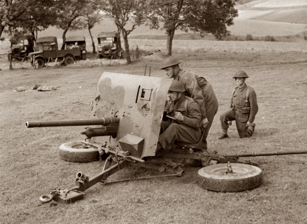 A black and white photo of a QF 2-pounder anti-tank gun in a firing position with its crew; one stands behind the gun while another sits at the ready, and a third crew member's role would be to supply ammunition, set against a backdrop of military vehicles and rolling hills.