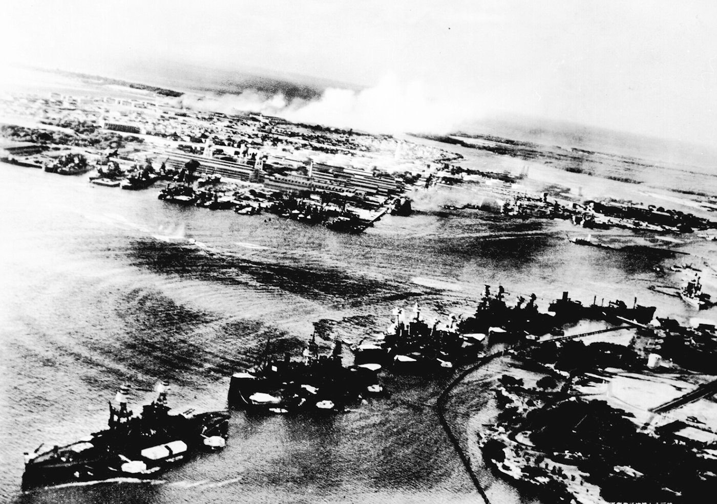 Aerial black and white photograph of the Japanese attack on Pearl Harbor from the perspective of the attackers on December 7, 1941. Smoke billows from Pearl Harbor, T.H., caught off guard during the aerial onslaught. The USS West Virginia is engulfed in flames amidst the chaos of the surprise assault. This image is a captured Japanese photograph from the historic event.