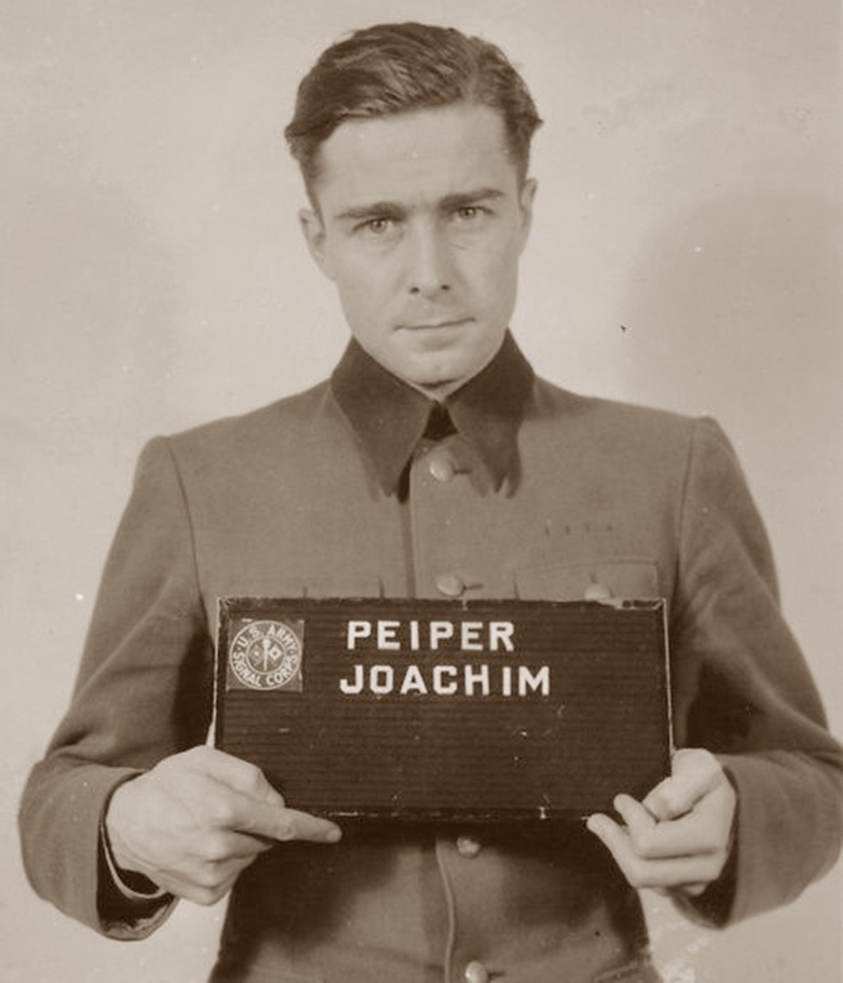 A historical mugshot of Joachim Peiper, taken after his arrest in 1944. He is seen in uniform, holding a nameplate with his name on it, displaying a somber expression.