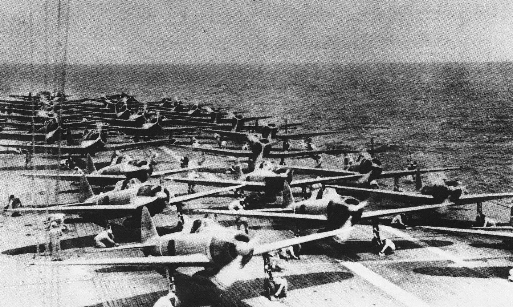 Historical black and white photo showing rows of Mitsubishi A6M2 Zero fighter aircraft lined up on the deck of the Imperial Japanese Navy aircraft carrier Shokaku, ready for the first strike wave against Pearl Harbor on December 7, 1941. The sea is visible in the background, indicating the carrier is at sea and poised for the imminent attack.