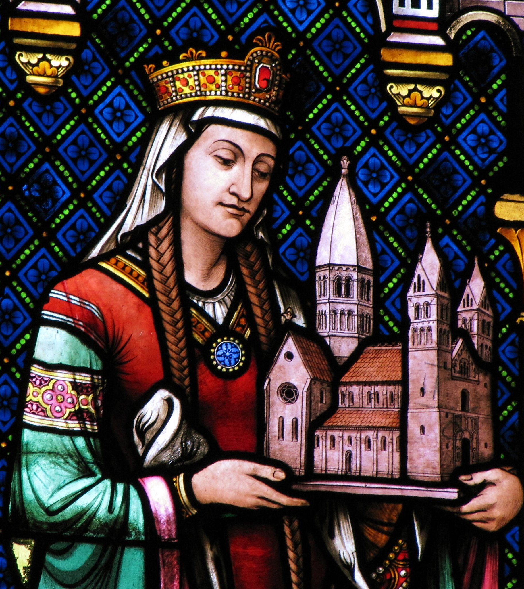 A stained glass portrait of Hildegard of Bingen at the Ste Foy Church in Conques, France. The image shows Hildegard as a serene, crowned figure, wearing a richly decorated red and green robe with a blue circular jewel at the center. She holds a model of a monastery, indicative of her role as a founder and leader of a religious community. The background features a blue, geometric pattern with fleur-de-lis motifs and elements of heraldry. The intricate details and vibrant colors of the stained glass illuminate her importance and veneration as a saint and a significant figure in the church. Her composed expression and the symbolic representation highlight her contributions to religious life and her status within the church.