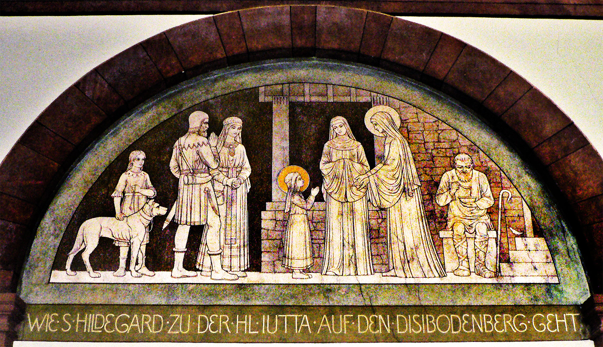 A semi-circular church tympanum featuring a relief depicting a young Hildegard of Bingen with St. Jutta. The scene shows a group of figures in medieval attire. In the center, a young girl, presumably Hildegard, stands before two veiled women, one of whom is likely St. Jutta, extending her hands in instruction or blessing towards Hildegard. The tympanum has a German inscription at the bottom, indicating the subject as Hildegard's journey to the Disibodenberg monastery.