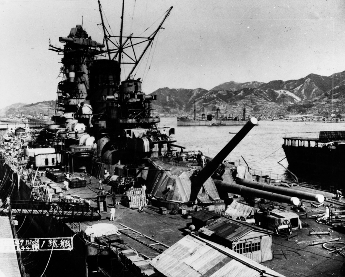 Black and white photo of the battleship Yamato during its construction in September 1941. In the background to the right is the carrier Hosho, and at the top of the image is the store ship Mamiya. The shipyard is bustling with activity, and the surrounding landscape features mountains. This historical photograph is cataloged by the U.S. Naval Historical Center with the number NH 63433.