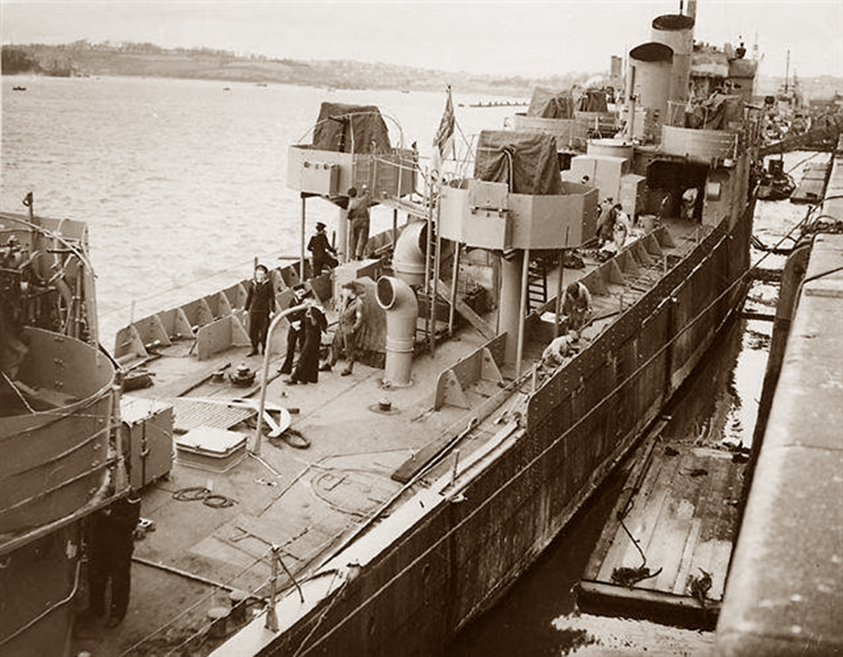 Historical black and white photograph showing the HMS Campbeltown during its conversion for the St. Nazaire Raid. The ship is docked and several figures can be seen working on its deck, which is outfitted with various equipment and structures, possibly as part of the modifications. The surrounding water is calm, and the opposite shore can be seen in the distance.