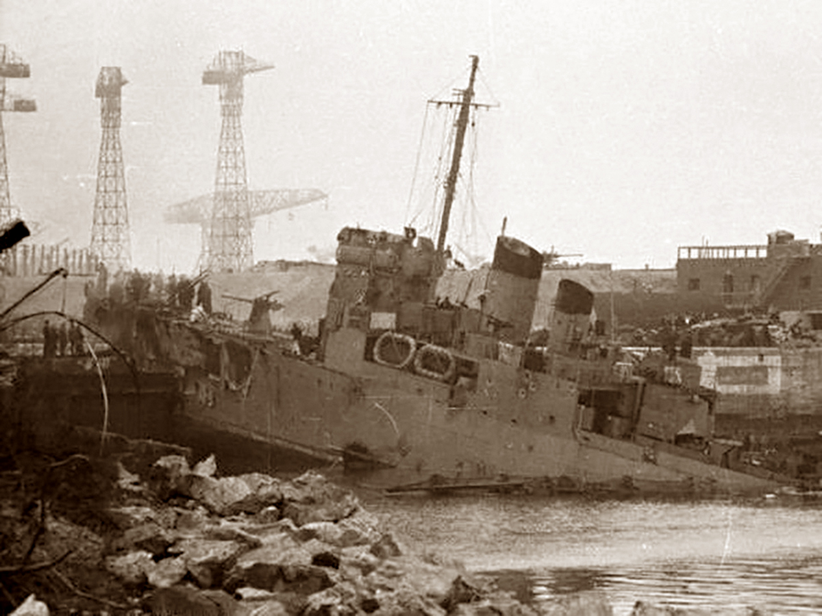 Black and white historical photo of the HMS Campbeltown lodged in the drydock at St. Nazaire, with visible damage and debris around it. The ship appears abandoned and partially destroyed. In the background, large skeletal tower structures, possibly cranes, loom over the scene, creating an atmosphere of post-battle desolation.