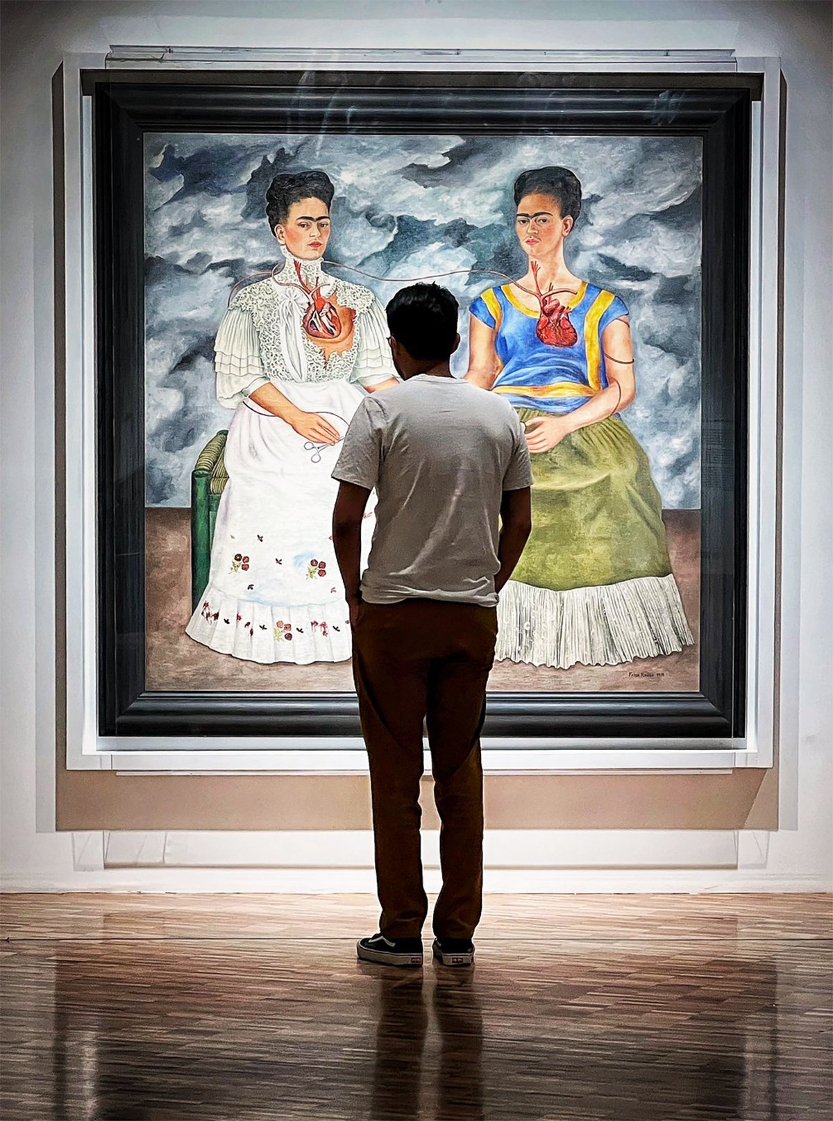 This image shows a person viewing "The Two Fridas" painting by Frida Kahlo on display at Museo de Arte Moderno in Mexico City. The viewer is standing with their back to the camera, providing a sense of scale to the artwork. The large painting features two self-portraits of Frida Kahlo sitting side by side against a stormy sky background. The person observing the painting is dressed casually in a white t-shirt and black pants, focusing intently on the details of the artwork.