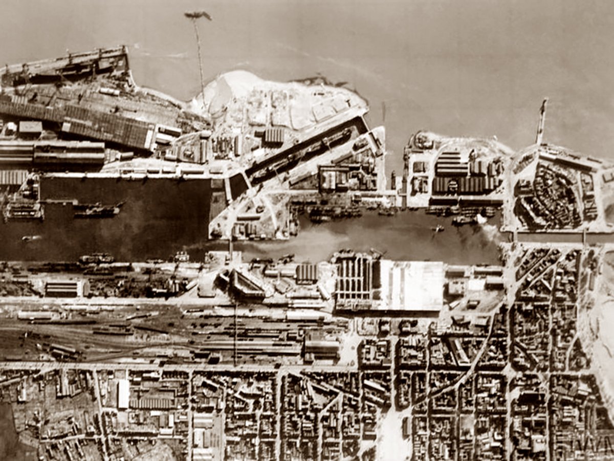 An aerial reconnaissance photograph showing the complex infrastructure of the docks at St. Nazaire prior to Operation Chariot. The image is detailed, displaying several docked ships, waterways, and the extensive dockyard facilities with various buildings, cranes, and piers. 