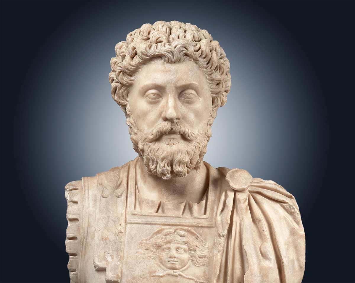 Bust of a bearded man, Marcus Aurelius (reign 161–180 CE), with detailed curly hair and beard. The sculpture displays intricate details, including a decorated chest armor with a medallion and draped cloak over one shoulder. The background is a gradient from dark to light grey, highlighting the figure.